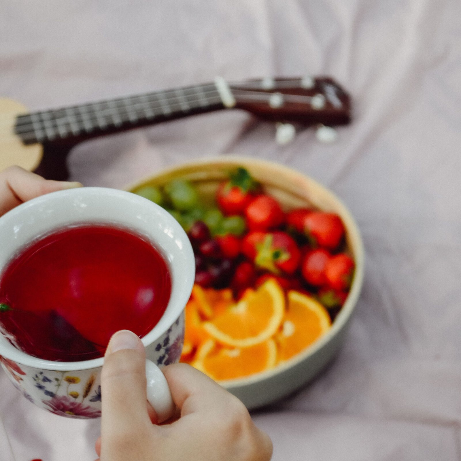 Vibrant bowl of fruits with herbal tea, hands holding cup near guitar.